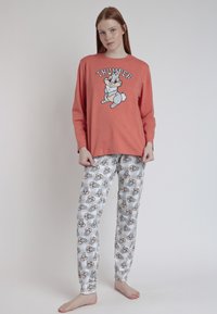 Mujer vestida con una blusa de manga larga color coral con un conejo de dibujos animados y pantalones de pijama blancos a juego con múltiples caras de conejo, de pie descalza sobre un fondo liso.