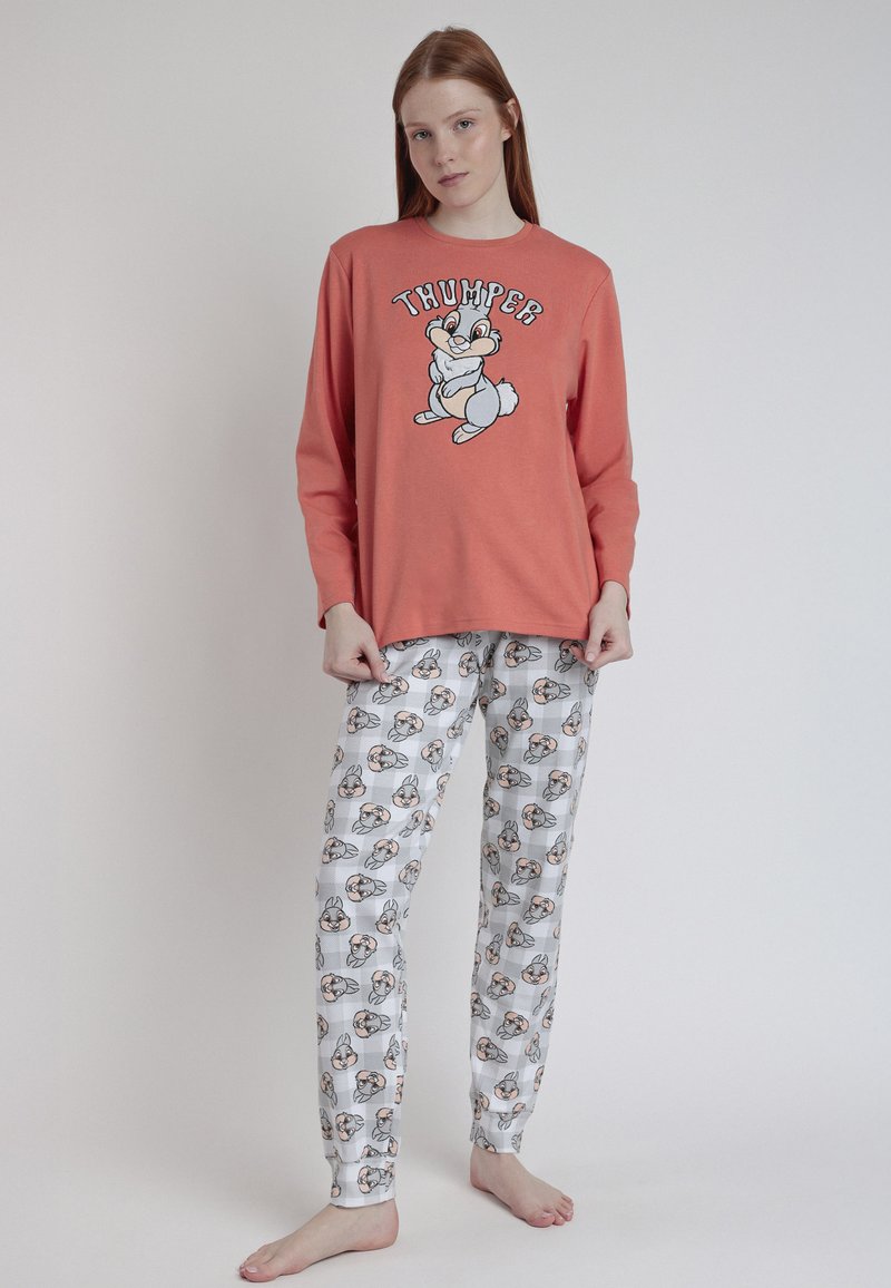 Mujer vestida con una blusa de manga larga color coral con un conejo de dibujos animados y pantalones de pijama blancos a juego con múltiples caras de conejo, de pie descalza sobre un fondo liso.