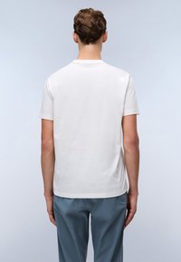 Tricou alb din bumbac cu mânecă scurtă și guler rotund, expus din spate. Material de bumbac neted, cu o croială obișnuită, asociat cu pantaloni albaștri deschiși.