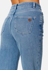 Jasnoniebieskie dżinsy charakteryzują się wysokim stanem, prostym krojem i dwiema tylnymi kieszeniami. Na prawej kieszeni widoczna jest mała brązowa skórzana metka z logo.
