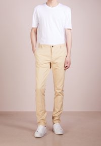 Béžové chinos s hladkou texturou, s předním zapínáním na knoflík, bočními kapsami a slim fit střihem, doplněné jednoduchým bílým tričkem a bílými teniskami.