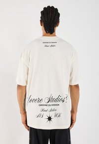 Cream-colored oversized t-shirt with short sleeves, featuring black text on the back, including brand name and design details.