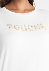 Hvid t-shirt med rund hals og korte ærmer, prydet med ordet "TOUCHÉ" i guld og perle-lignende dekorationer.