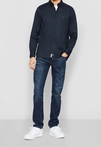 Man som står med en hand i fickan, klädd i en marinblå skjorta, blå jeans och vita sneakers mot en neutral bakgrund.