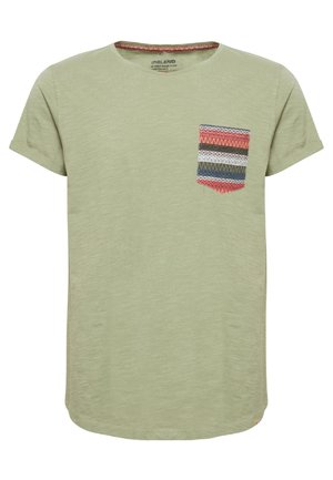 Homme marchant portant un t-shirt vert clair avec une poche poitrine à motif multicolore, un jean bleu et des baskets noires avec des lacets blancs.
