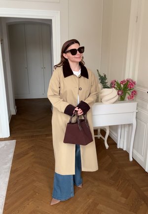 Woman wearing beige coat with dark collar and cuffs, blue wide-leg jeans, brown handbag, sunglasses, standing indoors near white table with flowers.