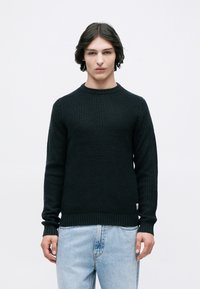 Νεαρός άνδρας με μεσαίου μήκους σκούρα μαλλιά, φορώντας ένα μαύρο υφασμάτινο πουλόβερ και ανοιχτόχρωμα μπλε τζιν, στέκεται μπροστά σε λευκό φόντο.