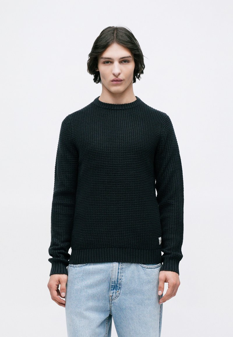 Νεαρός άνδρας με μεσαίου μήκους σκούρα μαλλιά, φορώντας ένα μαύρο υφασμάτινο πουλόβερ και ανοιχτόχρωμα μπλε τζιν, στέκεται μπροστά σε λευκό φόντο.