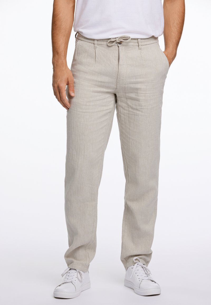 Homme portant un pantalon en lin beige avec une taille à cordon et des baskets blanches, debout avec une main dans la poche.