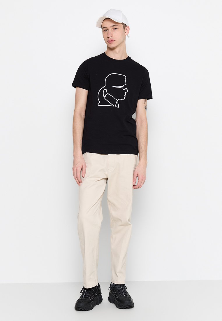 Camiseta de algodón negra con un diseño de perfil con contorno blanco. Combinada con pantalones beige y zapatillas negras de textura. Gorra en la cabeza.