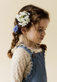 Dziecko w profilu bocznym, z warkoczami ozdobionymi białymi i niebieskimi kwiatami. Ma na sobie beżowy top w wzorki oraz niebieski dżinsowy ogrodnik z falbankami.