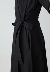 Robe noire en tissu élégant, avec une taille cintrée ornée d'un nœud et une jupe fluide. Design simple et minimaliste avec une couleur unie.