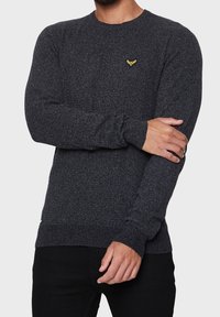 Pull en maille gris foncé à manches longues. Doté d'un col rond et d'un petit emblème jaune sur la poitrine. Le tissu semble texturé.