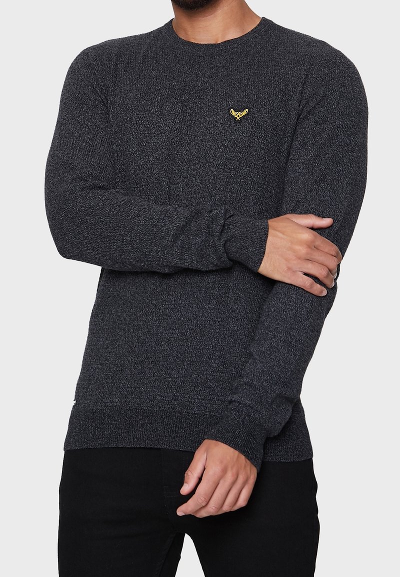 Pull en maille gris foncé à manches longues. Doté d'un col rond et d'un petit emblème jaune sur la poitrine. Le tissu semble texturé.