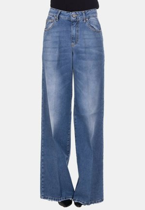 Donna che indossa jeans a vita alta, sbiaditi blu a gamba larga abbinati a un top nero e scarpe nere, in piedi davanti a uno sfondo chiaro e uniforme.