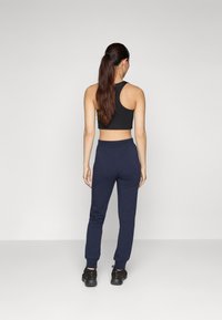 Schwarzes, kurzes Tanktop in Kombination mit navy-farbenen, taillierten Jogginghosen, die elastische Bündchen und Seitentaschen haben. Das Modell trägt schwarze Sportschuhe.