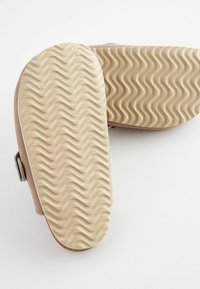 Ljusbruna slip-in-skor med en texturerad gummisula som har ett vågigt mönster, som visar en slät ovandel och en sidospänne-detalj.