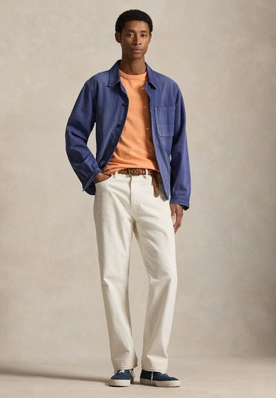 Νεαρός άνδρας με τα χέρια στις τσέπες, φοράει μπλε ναυτικό μπουφάν, πορτοκαλί πουκάμισο, άσπρο παντελόνι με πλεγμένη ζώνη και μπλε αθλητικά, στέκεται σε ουδέτερο φόντο.