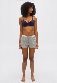 Γυναίκα με σγουρά μαλλιά που φοράει σκούρο μπλε σουτιέν και γκρι σορτς Polo Ralph Lauren, όρθια ξυπόλυτη και κοιτάζει προς τα αριστερά της, μπροστά σε απλό φόντο.