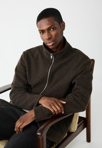 Tmavohnedá bunda na zips s vysokým golierom a jemnou textúrou, oblečená na šedej blúzke. Model sedí na drevenej kresle so založenými rukami.