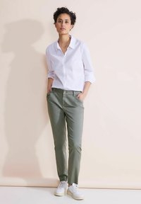 Femme debout contre un mur beige, portant une chemise blanche boutonnée, un pantalon vert olive, des baskets blanches, les mains dans les poches, et une expression neutre.