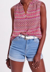 Haut sans manches en v sur le devant avec un motif géométrique multicolore en rose, blanc et jaune, associé à un short en denim bleu clair et une ceinture blanche.