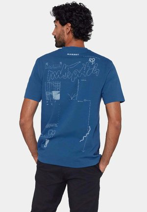 Hombre con cabello rizado que lleva una camiseta azul de Mammut con diseños abstractos en líneas blancas y texto en la espalda, manos en los bolsillos, de espaldas.