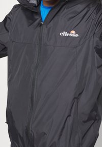 Veste imperméable noire avec fermeture éclair sur le devant, capuche et logo. Présente une texture lisse et un léger éclat, avec deux poches latérales.