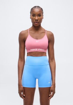 Kobieta stojąca zwrócona przodem, ubrana w różowy sportowy biustonosz Nike Pro i wysokowalne niebieskie spodenki Nike na jednolitym tle.