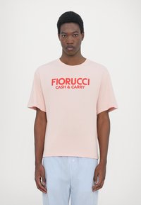Ανοιχτό ροζ κοντομάνικο μπλουζάκι φτιαγμένο από βαμβάκι, με μια έντονη κόκκινη γραφική εκτύπωση που λέει "FIORUCCI CASH & CARRY" στο μπροστινό μέρος.