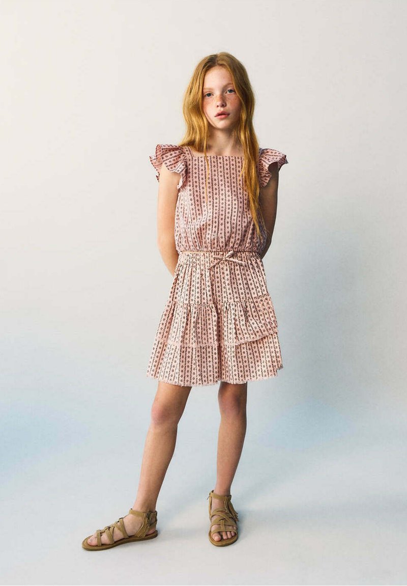 Jeune fille aux longs cheveux roux portant une robe rose à motifs et des sandales marron, debout devant un fond clair uni.