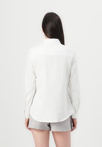 Chemise blanche à manches longues avec un empiècement au dos, ourlet arrondi et détails en boutons. Le matériau semble lisse et léger. Assortie avec un short gris.