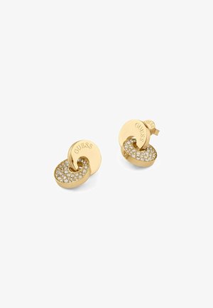 Boucles d'oreilles en or avec un design torsadé, une surface texturée et des cristaux transparents scintillants incrustés dans la partie inférieure. Marque visible 'GUESS'.