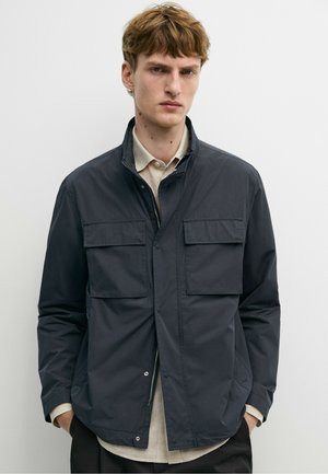 Junger Mann mit kurzen braunen Haaren, der eine dunkelblaue Jacke über einem beigen Hemd trägt und vor einem schlichten weißen Hintergrund steht.