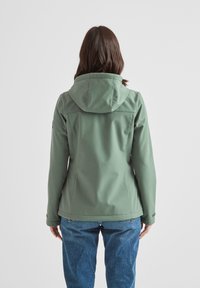 Green softshell jacket with a hood, fitted silhouette, zippered pockets, and a smooth texture. Worn over blue denim jeans.