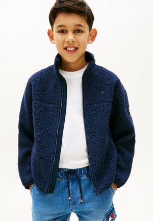 Junge lächelt, trägt eine marineblaue Fleecejacke über einem weißen Hemd und blaue Jeanshosen, die Hände in den Taschen, vor einem einfarbigen Hintergrund.