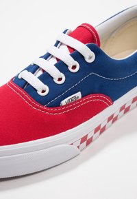 Rode en blauwe canvas sneakers met witte veters. Heeft een geruit patroon in wit en rood op de zool. Het Vans-logo is zichtbaar aan de zijkant.