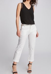 Top senza maniche nero con spalline in pizzo, abbinato a pantaloni bianchi con risvolto e décolleté neri con cinturino alla caviglia. Accenti di cintura argentata.