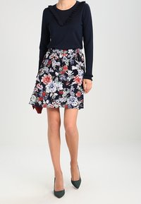 Mörkblå långärmad topp med volangkrage, matchad med en blommig kjol i mörkblått, rött och vitt. Bärs med gröna klackar.