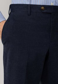 Uomo che indossa pantaloni a quadri blu scuro con tasche laterali e una camicia a righe blu infilata dentro, con una mano in tasca.
