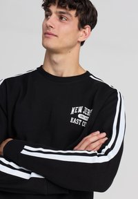 Felpa nera con strisce bianche sulle maniche, con grafica testuale frontale. Collo e polsini a coste, tessuto morbido, vestibilità rilassata.