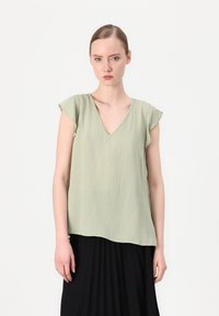 Lys grønn v-hals skjorte med korte cap-ermer og et litt teksturert stoff, kombinert med et svart plisse skjørt. Enkelt design.