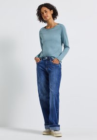 Žena s kudrnatými vlasy, oblečená v světle modrém dlouhém rukávu, volných modrých džínech s manžetami a béžových teniskách, stojící s rukama v kapsách.