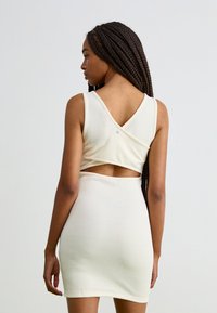 Rochie de culoare crem, dintr-un material texturat, cu un decolteu adânc la spate și detaliu decupat în talie. Designul fără mâneci subliniază silueta.