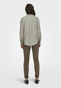 Camisa de manga larga gris claro con un pliegue en la espalda, combinada con pantalones ajustados de ante marrón y sandalias negras de tacón. Diseño minimalista, ajuste a medida.