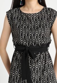 Černé šaty se geometrickým řetězcem vzoru v bílé. Krátké rukávy, pásek v pase a hladká textura.
