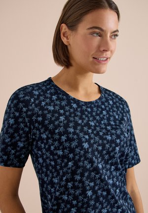 Femme aux cheveux bruns courts portant une chemise bleu marine à motifs de petits palmiers bleu clair, souriante et regardant sur le côté.