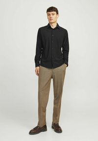 Camicia nera a maniche lunghe con bottoni abbinata a pantaloni a quadri marroni chiari e scarpe marroni scure, in piedi contro uno sfondo neutro.