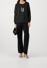 Zwart langemouw sweatshirt met een grafisch logo, gecombineerd met hoge zwarte jeans en witte sandaaltjes met bandjes, terwijl een zwarte clutch wordt vastgehouden.