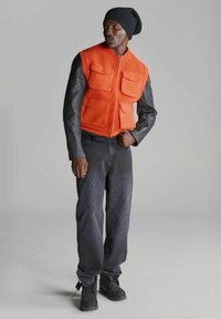 Gilet arancione lavorato a maglia con quattro tasche frontali, maniche in pelle nera, jeans neri con motivo a diamante e calzature nere.
